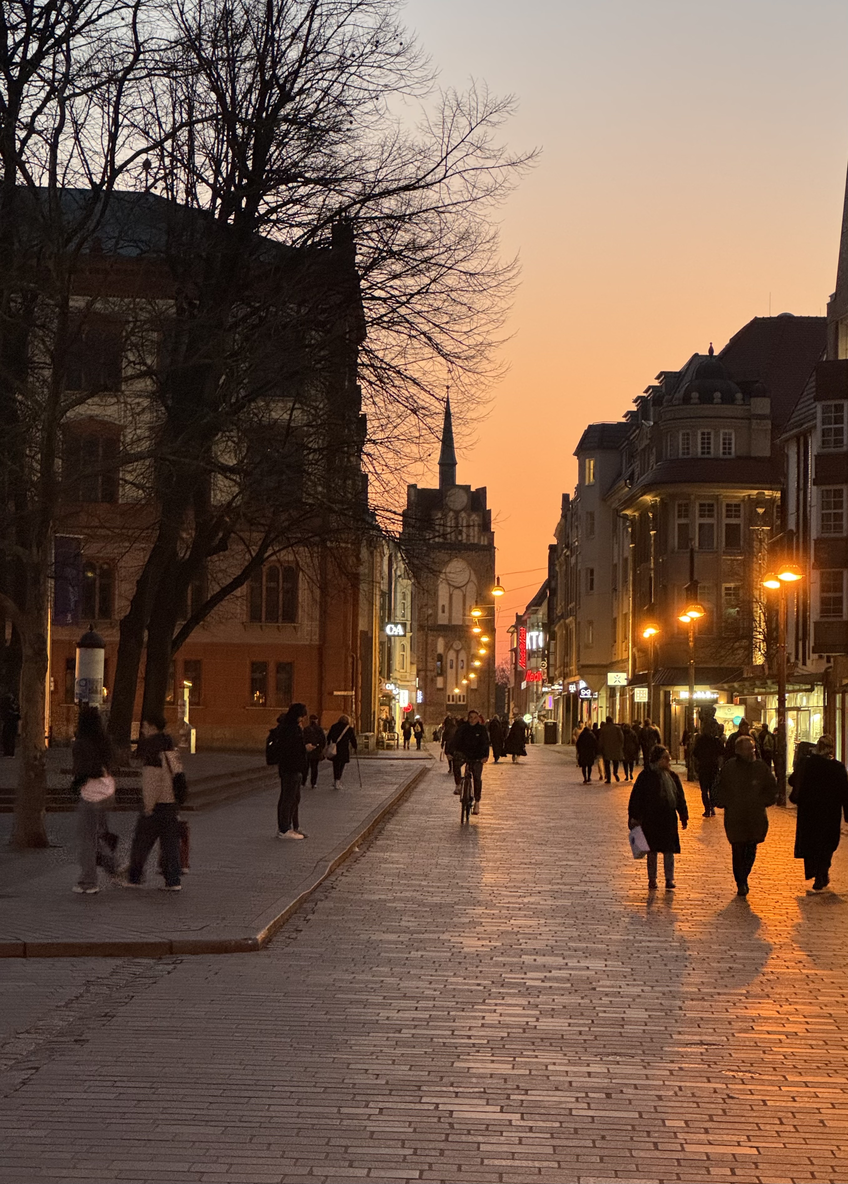 Eine belebte Straße in einer Stadt während des Sonnenuntergangs. Der Himmel ist in warmen Orangetönen gefärbt. Auf der linken Seite sind große, alte Gebäude und kahle Bäume zu sehen. Menschen spazieren und ein Fahrradfahrer fährt auf der Straße. Rechts sind Geschäfte mit leuchtenden Schildern und Laternen, die die Straße beleuchten. Im Hintergrund ist ein Gebäude mit einem Uhrturm zu erkennen. Die Atmosphäre wirkt ruhig und stimmungsvoll.